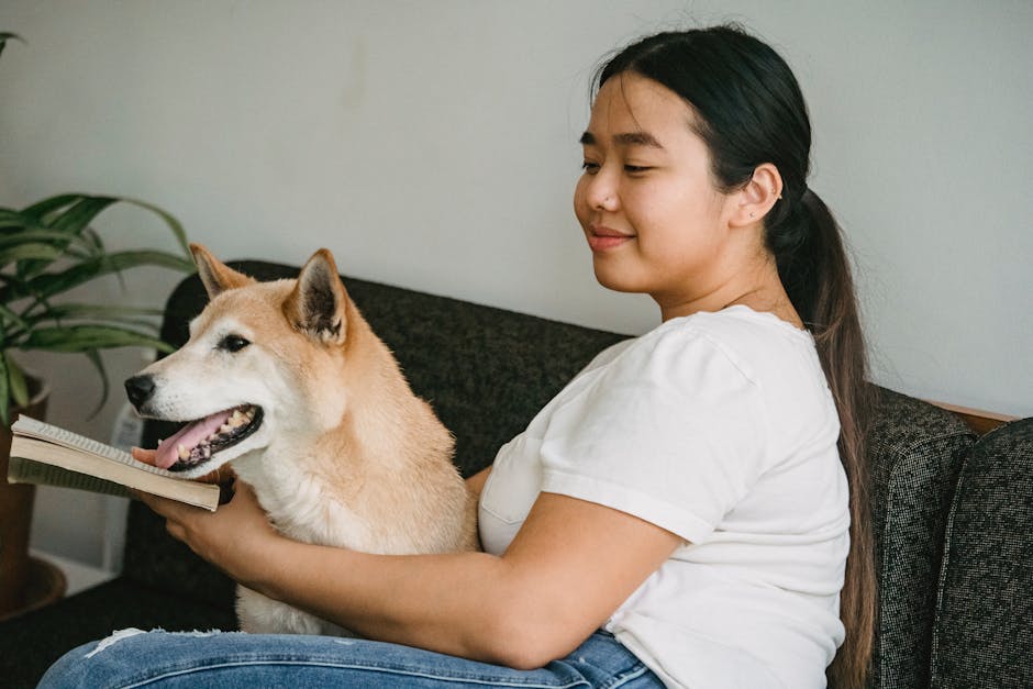 Side view of cheerful ethnic female resting on sofa and reading novel while cute Shiba inu putting muzzle on book